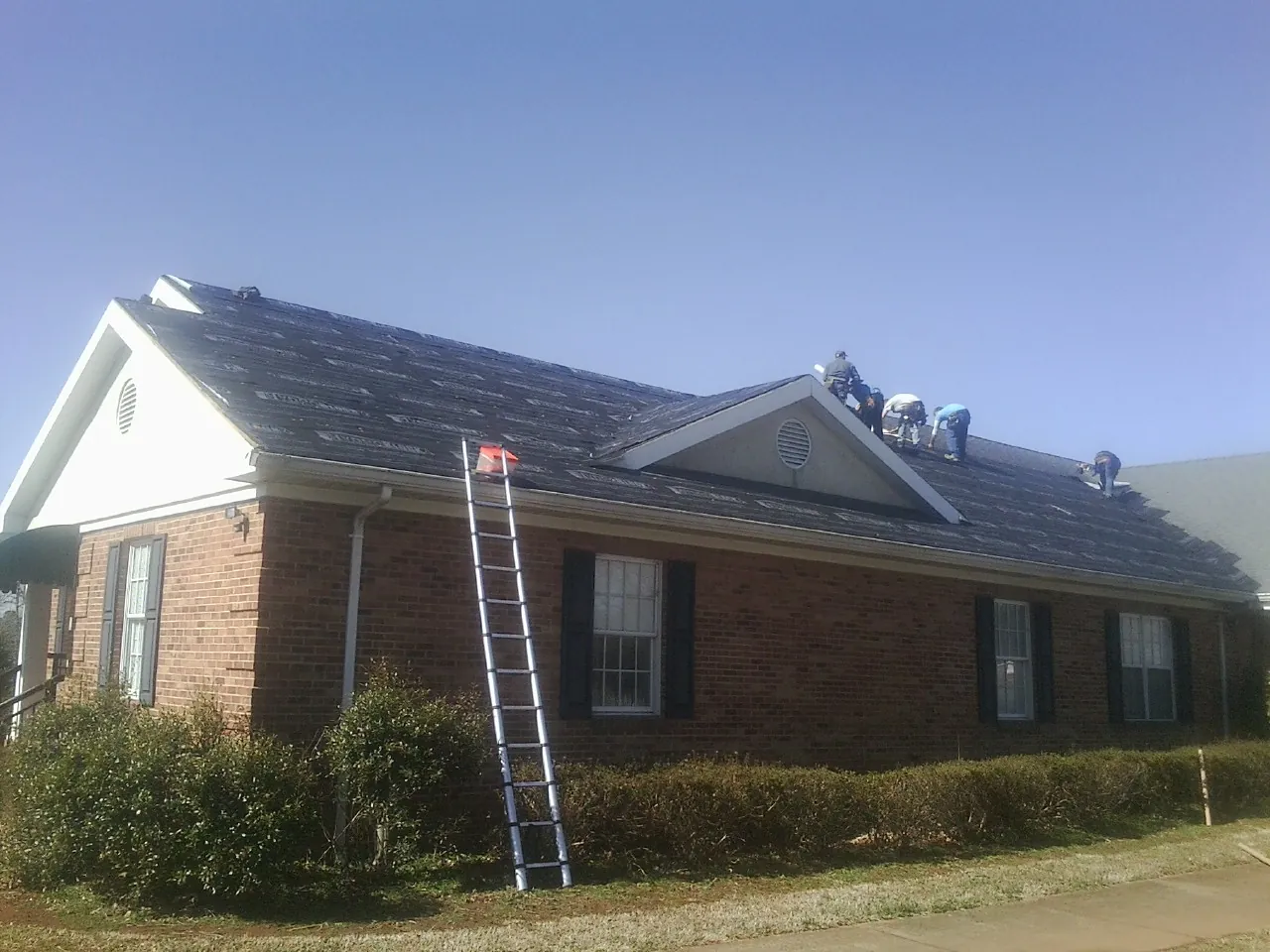 Crew tearing shingles off a brick ranch roof with a ladder set — Emergency Roof Repair work by our crew in South Otselic