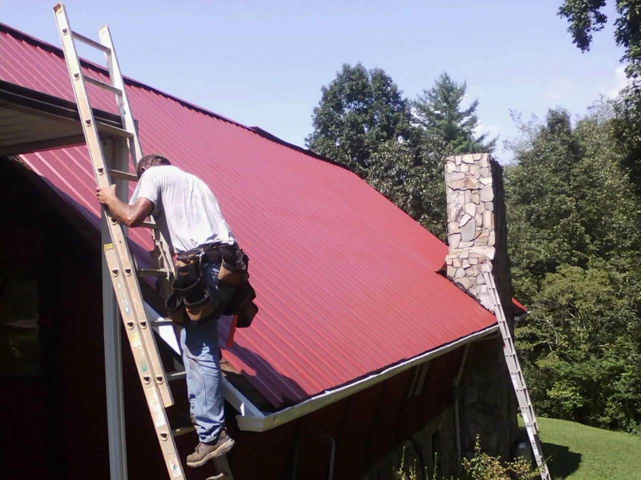 Roofer on a ladder beside a red metal roof and stone chimney — Soffit & Fascia Repair by Roofing Brothers in South Otselic